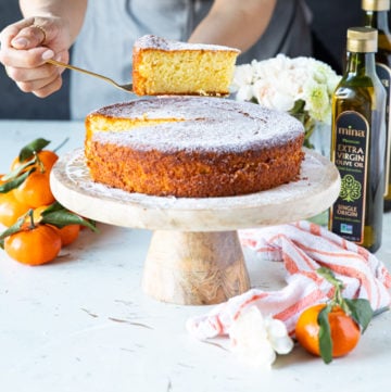 A hand holding a slice of olive oil cake over the serving olate with some clementine oranges and a tea towel around it