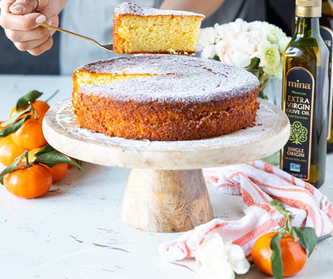 A hand holding a slice of olive oil cake over the serving olate with some clementine oranges and a tea towel around it