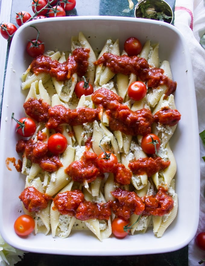Assembly of ricotta stuffed shells recipe in dish, with tomatoes and marinara sauce on top.