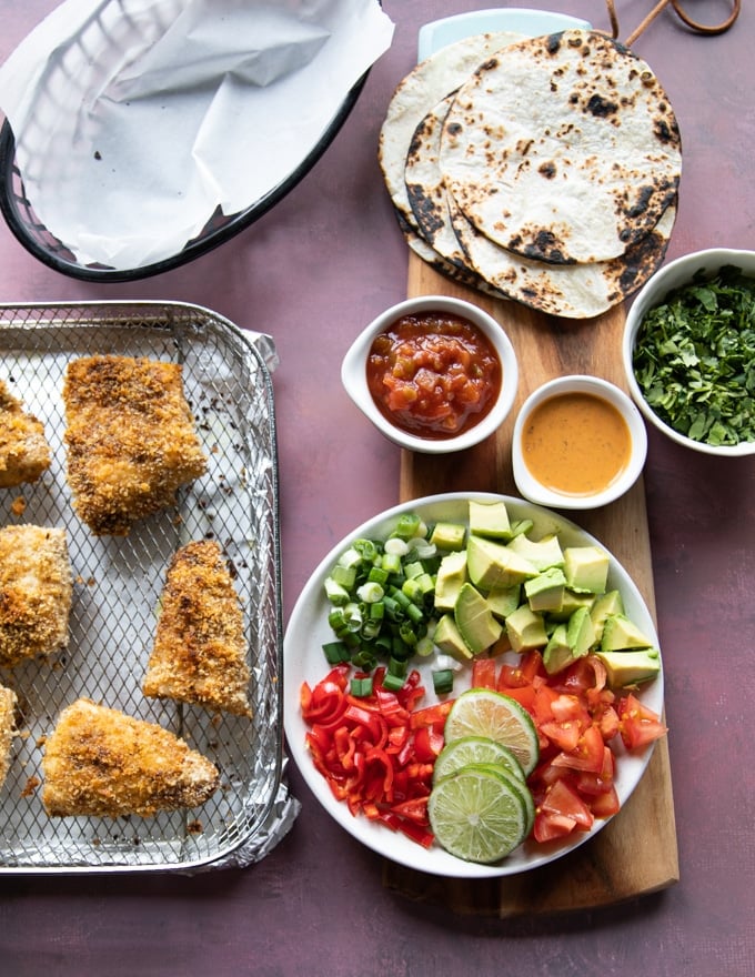 ingredients for air fryer fish and to make air fryer fish tacos including tortillas charred, the ready fish, some chopped avocados, chipotle sauce, salsa, fresh cilantro and lime