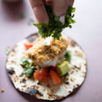 a flour tortilla with toppings like avocados, tomatoes, cilantro and then the air fryer fish is placed ontop and a hand is sprinkling more cilantro over the top