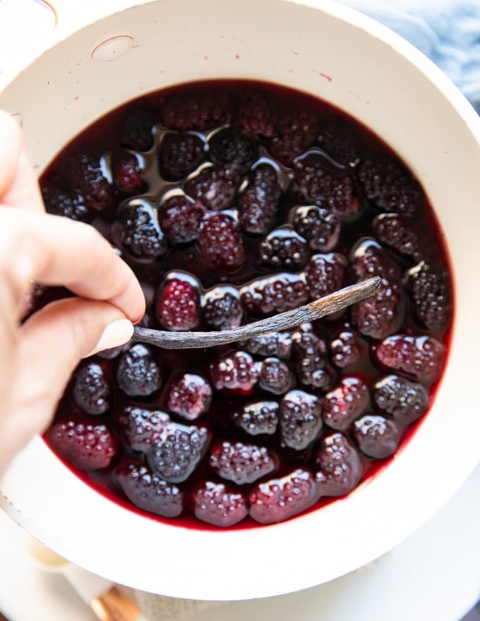 A hand holding a vanilla bean adding it to the pot of simmering marionberry for the blackberry jam recipe