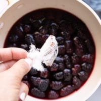 A hand throwing the cheesecloth with the whole spice wrapped right into the pan of simmering blackberry jam