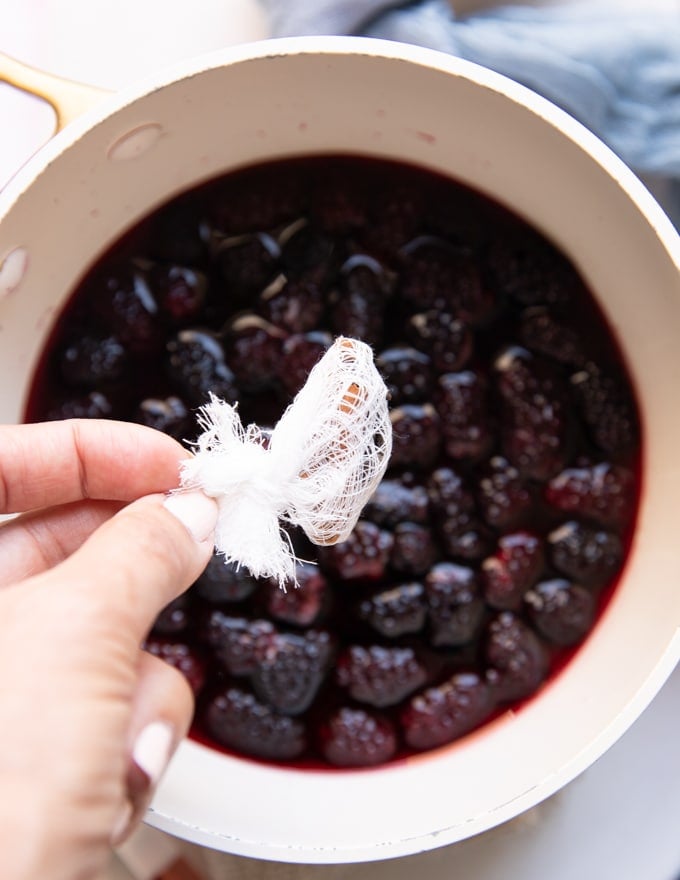 A hand throwing the cheesecloth with the whole spice wrapped right into the pan of simmering blackberry jam