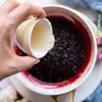 A hand pouring off the gelatin mixture into the pot of simmering jam once blackberry jam recipe is finished to thicken it
