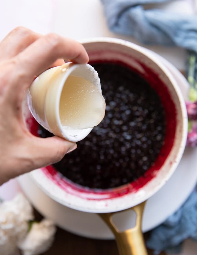 A hand pouring off the gelatin mixture into the pot of simmering jam once blackberry jam recipe is finished to thicken it