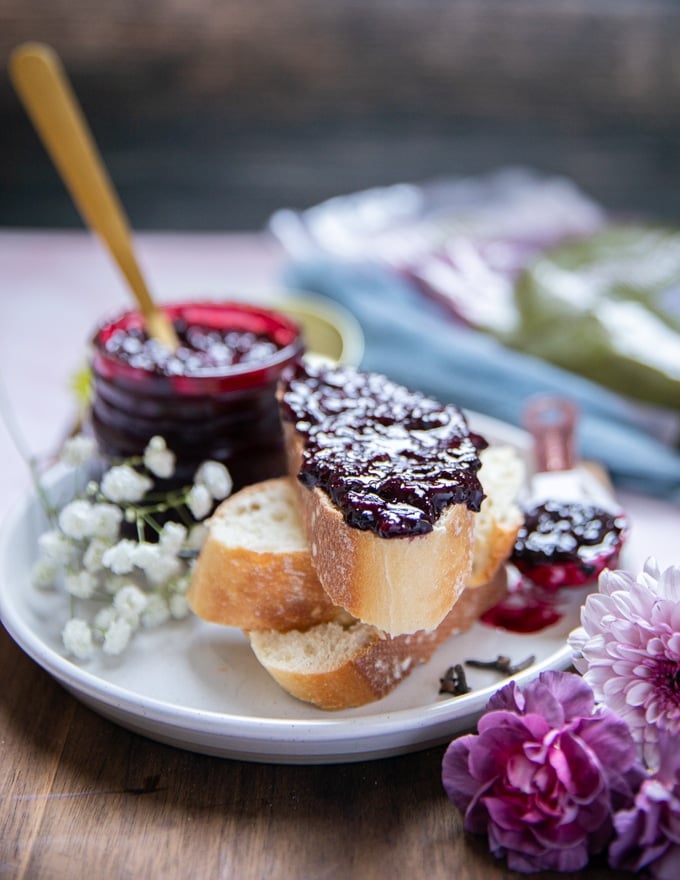 A baguette toast spread with blackberry jam on a plate with a jar of blackberry jam recipe in the background on a white plate and a purple flower on the table. 
