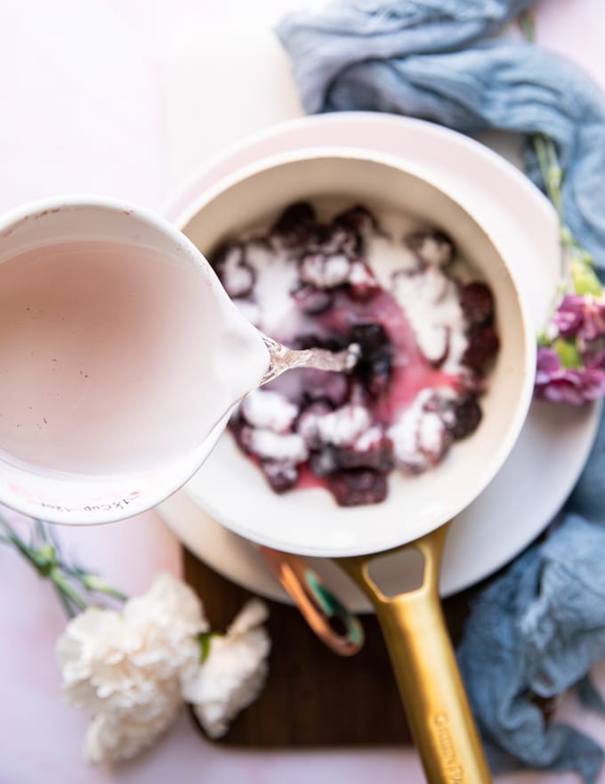 A bowl of water pouring into a pot with the marionberry blackberry and sugar ready to simmer