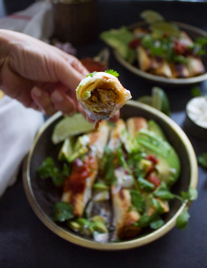 A hand holding a chicken flautas bitten to show the cheesy chicken filling and the crunchy tortilla wrap