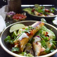 A plate of chicken flautas layerd over each other drenched in toppings like avocadoes, salsa, cilantro, sour cream and lime wedges