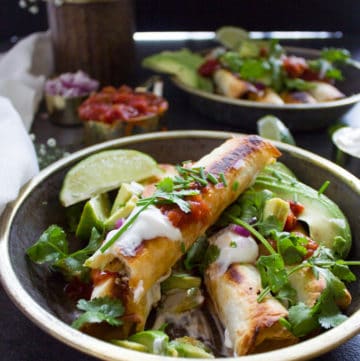 A plate of chicken flautas layerd over each other drenched in toppings like avocadoes, salsa, cilantro, sour cream and lime wedges