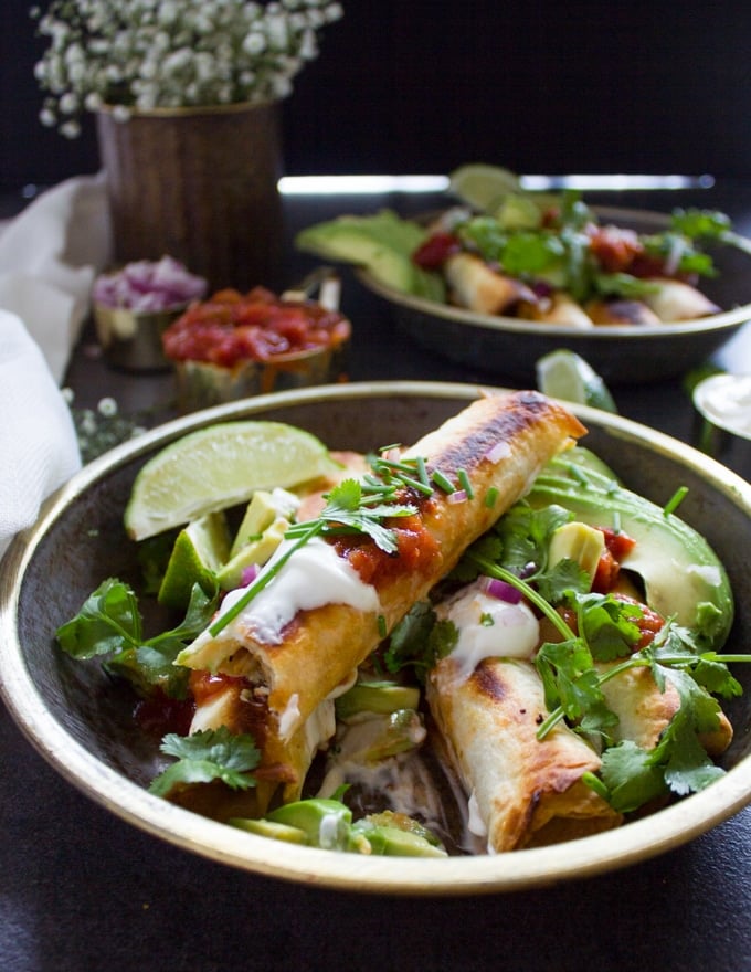 A plate of chicken flautas layerd over each other drenched in toppings like avocadoes, salsa, cilantro, sour cream and lime wedges
