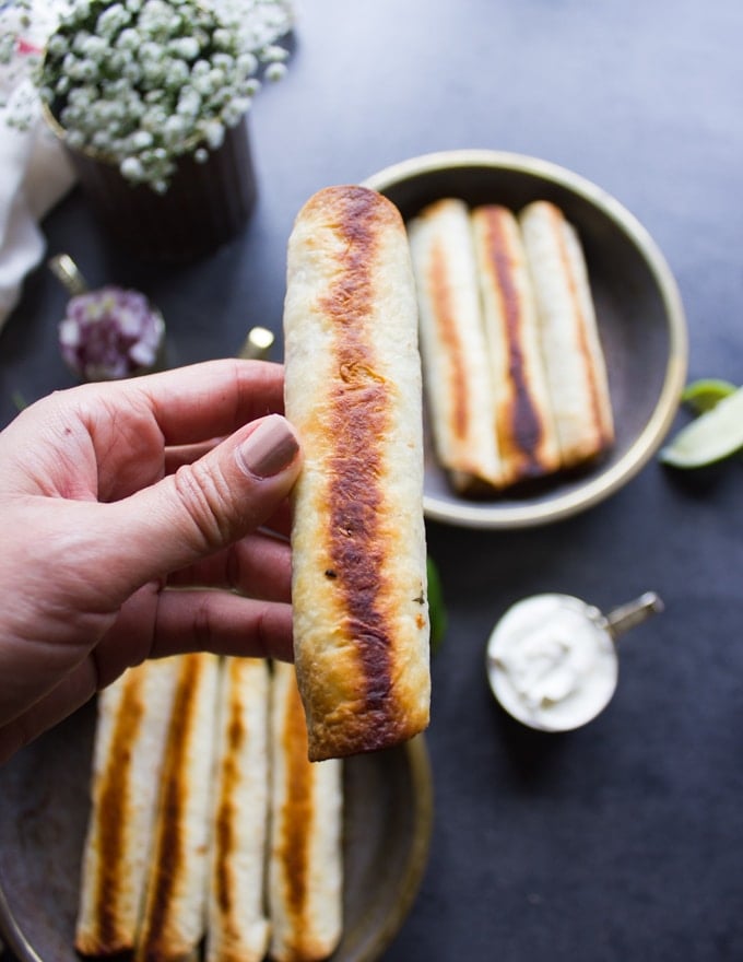 A hand holding a golden crisp chicken flautas out of the oven showing how crisp and golden it is