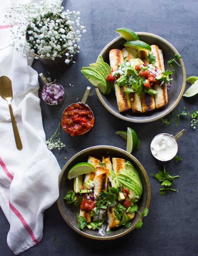 2 plates of chicken flautas surrounded by different toppings to add on such as a bowl of salsa, a bowl of guacamole, a plate of chopped onions, chopped avocados, a bowl of sour cream, fresh cilantro, lime wedges