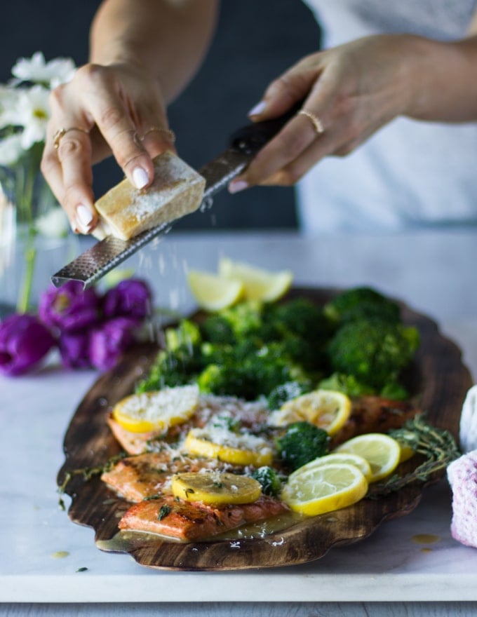 A hand grating some parmesan cheese over the top of the sockeye salmon recipe plated on a plate served with broccoli