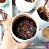 a hand holding the brownie in a mug right out of the microwave showing the cooked brownies and melted chocolate chips on top