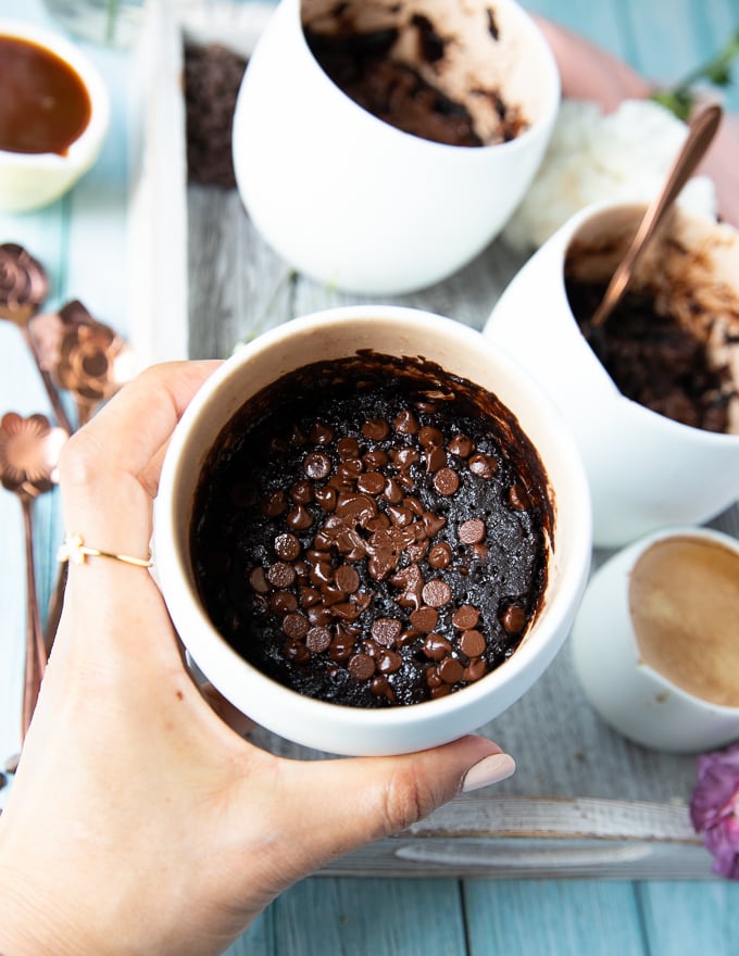 a hand holding the brownie in a mug right out of the microwave showing the cooked brownies and melted chocolate chips on top