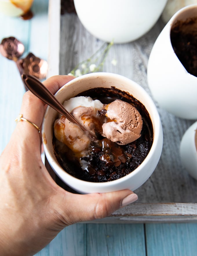 A hand holding a brownie in a mug loaded with scoops of chocolate and vanila ice cream and caramel sauce