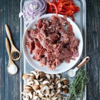 A tray with thinly sliced meat, thinly sliced onion, peppers ad mushrooms. There are spoons of wslices on the side such as spoon of salt, spoon of pepper and spoon of onion powder and fresh herbs