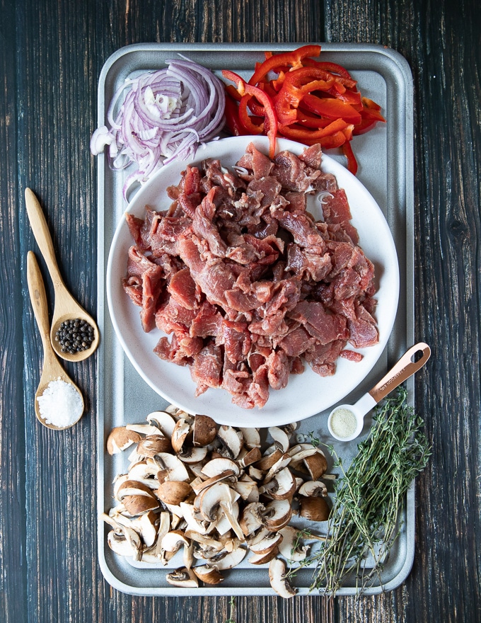 A tray with thinly sliced meat, thinly sliced onion, peppers ad mushrooms. There are spoons of wslices on the side such as spoon of salt, spoon of pepper and spoon of onion powder and fresh herbs