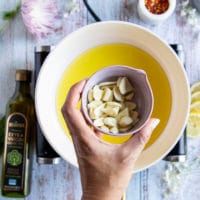 A hand holding the sliced garlic over a skillet of olive oil