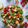 A plate of caprese salad with basil leaves surrounding it and homemade croutons on a white board with cherry tomatoes around it