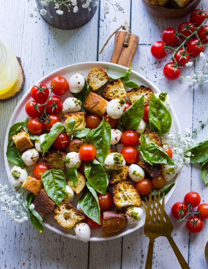 A plate of caprese salad with basil leaves surrounding it and homemade croutons on a white board with cherry tomatoes around it
