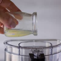 A hand drizzling some lemon juice through the feed tube of a food processor