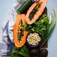 A papaya cut in half over a wooden board showing the beauty of this fruit surrounded by a bunch of cilantro, scallions and a cucumber