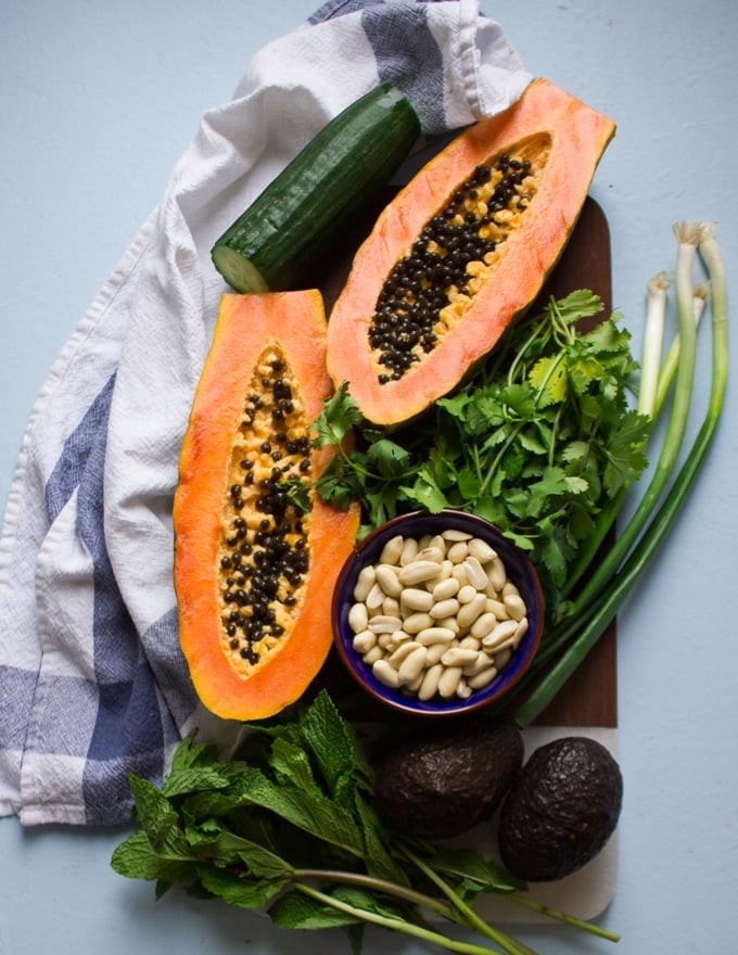 A papaya cut in half over a wooden board showing the beauty of this fruit surrounded by a bunch of cilantro, scallions and a cucumber