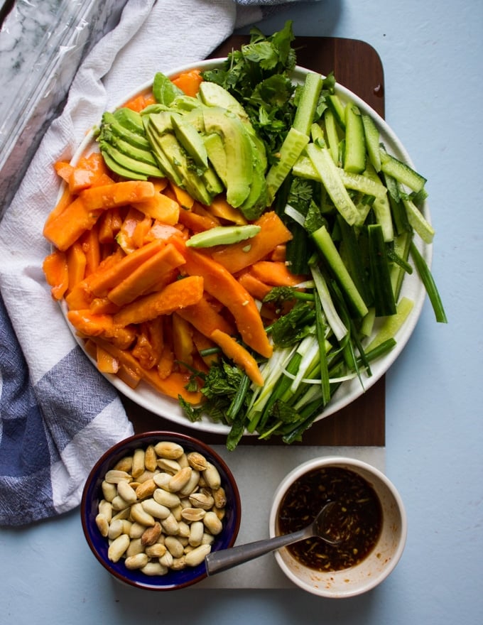 papaya salad recipe ingredients in a bowl including papaya, avocados, cucumbers, scallions, fresh mint, cilantro, a bowl with papaya salad dressing and a bowl of cashews