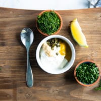 A bowl with tartar sauce ingredients like mayo, sour cream, mustard, capers, horseradish, two bowls of herbs (parsley and dill) and a lemon wedge.