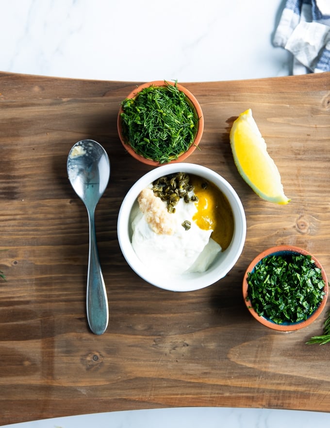 A bowl with tartar sauce ingredients like mayo, sour cream, mustard, capers, horseradish, two bowls of herbs (parsley and dill) and a lemon wedge.