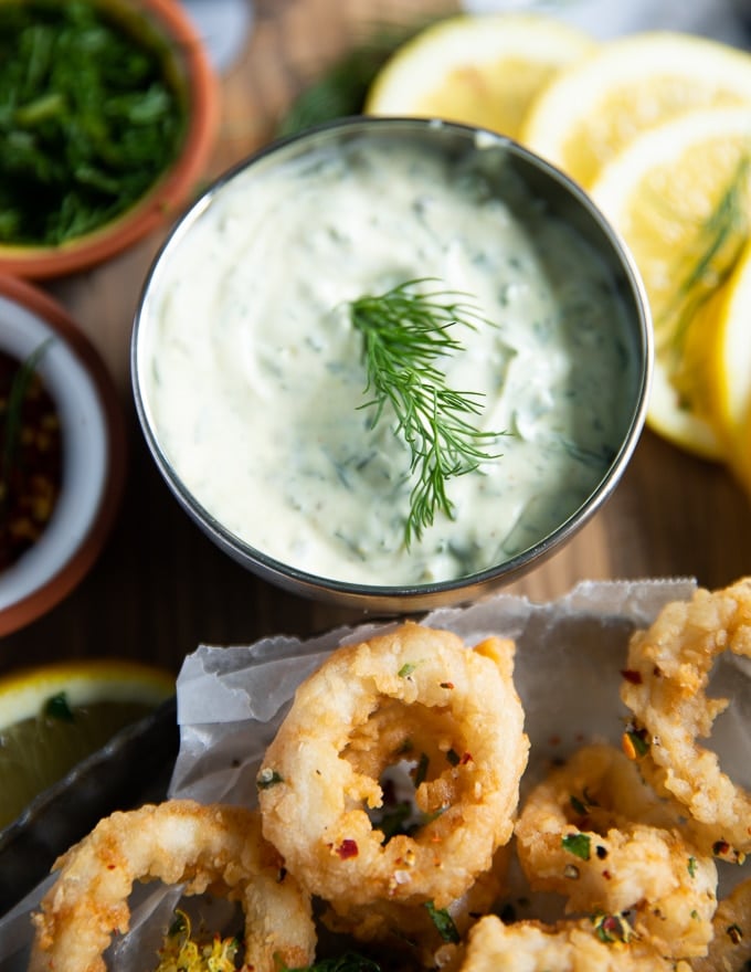 A bowl with tartar sauce and with a fresh dill sprig served close to a plate of calamari