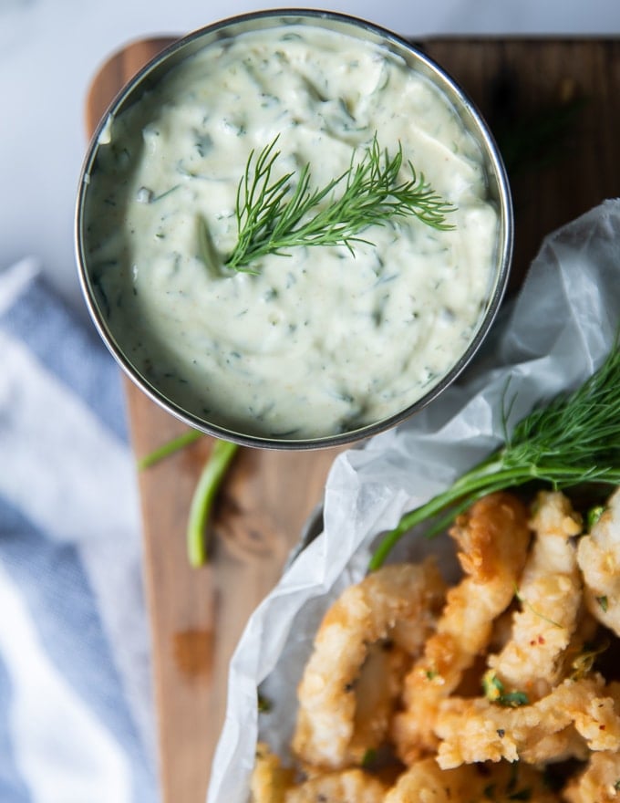 A bowl of tartar sauce with a sprig of dill on the top placed on a wooden board with some fresh dill and lemon around it