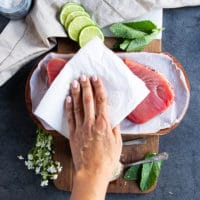 A hand holding a kitchen towel patting down the tuna steaks dry before seasoning