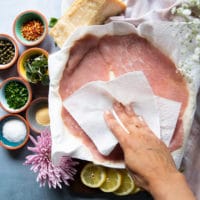 Thin slices of veal scallopini on a plate and a hand patting dry the veal using a paper towel