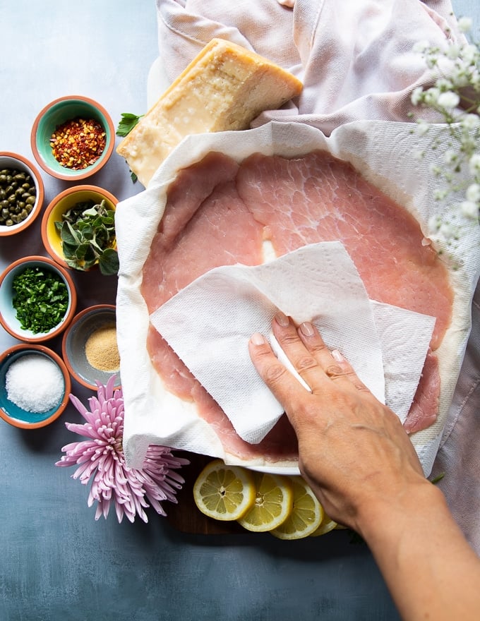 Thin slices of veal scallopini on a plate and a hand patting dry the veal using a paper towel 