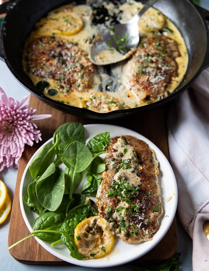 A plate with a serving of veal scallopini next to some spinach salad on a wooden board