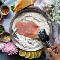 A hand holding a slice of veal scallopini using a tong and dredging it in the flour bowl