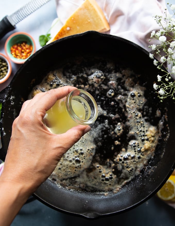 melted butter and garlic cooking in the same cast iron pan and a hand pouring some lemon juice to make the piccata sauce