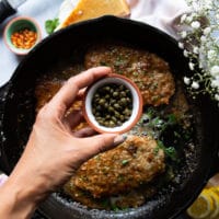 A hand adding in some capers to the piccata sauce