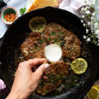 A hand finishing off the piccata sauce with a touch of cream in a small cup and the veal being added back to the pan