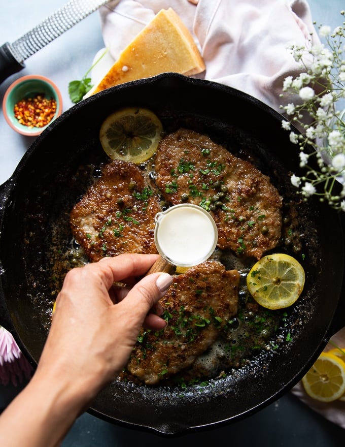 A hand finishing off the piccata sauce with a touch of cream in a small cup and the veal being added back to the pan