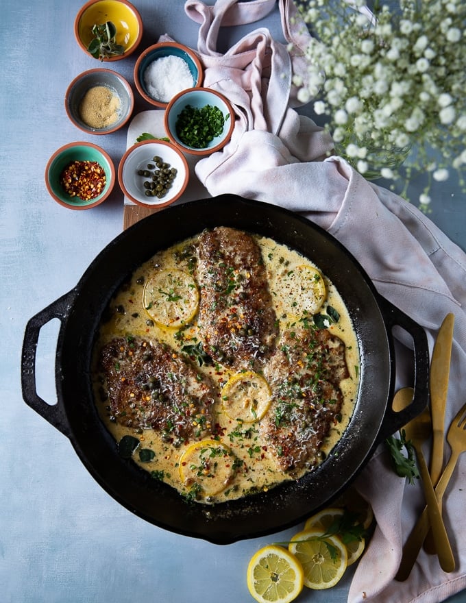 the veal scallopini in a cast iron skillet with the piccata sauce and surrounded bu extra herbs, chilli flakes and parmesan cheese for garnish if needed