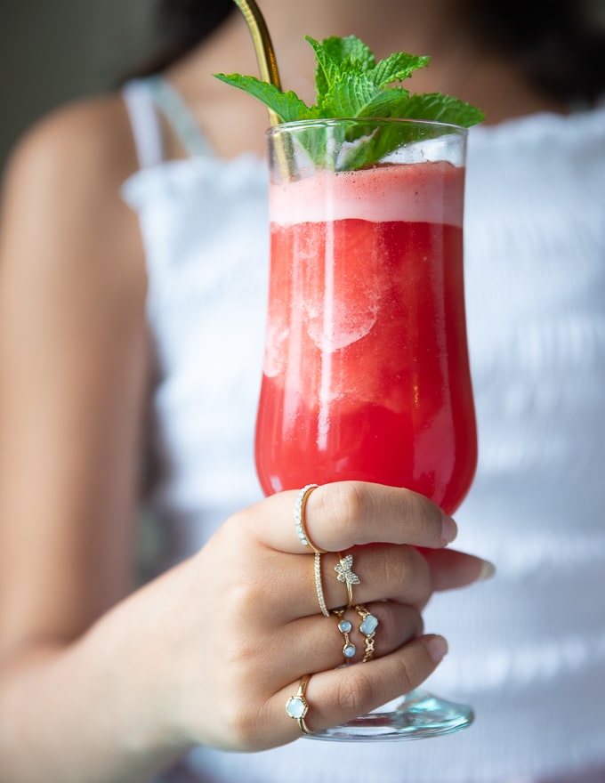 A hand holding a tall cup of watermelon slushie with a mint sprig on top