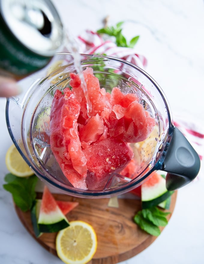 A hand pouring some soda drink into the blender with the frozen watermelon to blend into a slushie drink