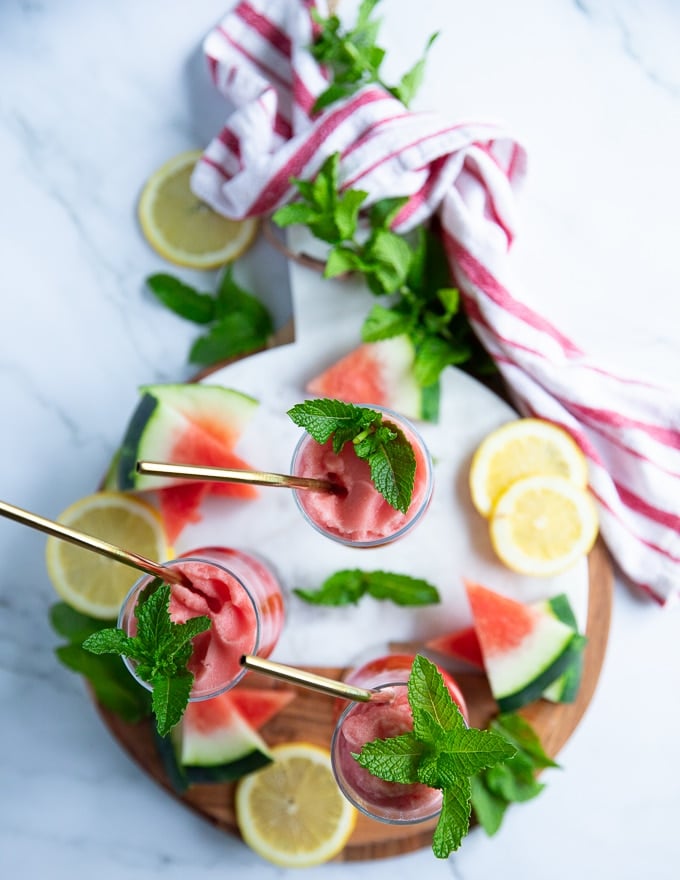 A wooden board with three cups of watermelon slushie and some watermelon wedges, lemon slices and fresh mint around it