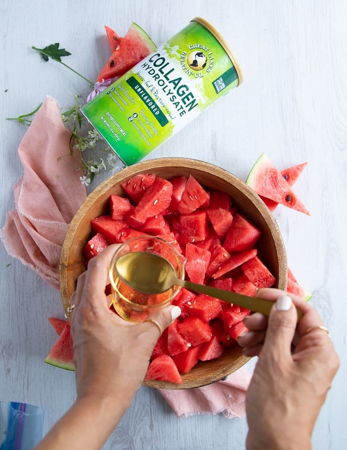 a lage bowl of cubed watermelon and a hand adding the simple syrup to the watermelon