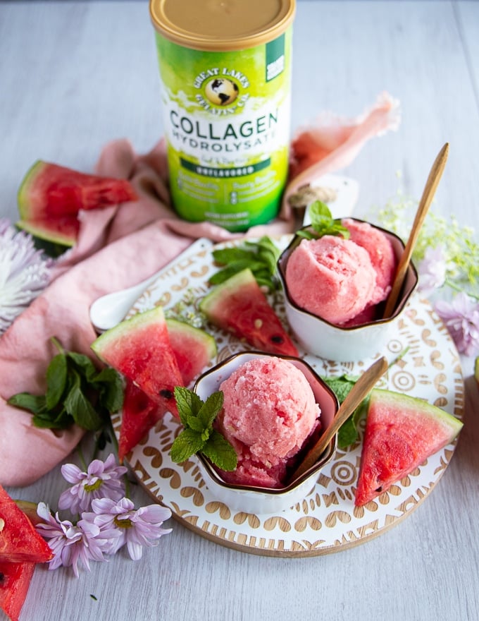 two bowls of watermelon sorbet on a white board with watermelon wedges on the board and mint leaves, and a pink kitchen towel